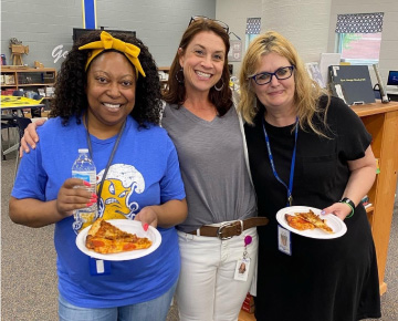 Three teachers standing in school cafeteria with pizza on paper plates
